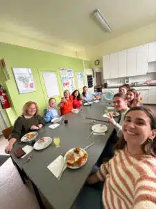 Gezellige lunchscène in de klas waar studenten en een leraar genieten van een maaltijd naast het logo "Deze school duikt in taal", uitstralend gemeenschapsgevoel en schoolcultuur.